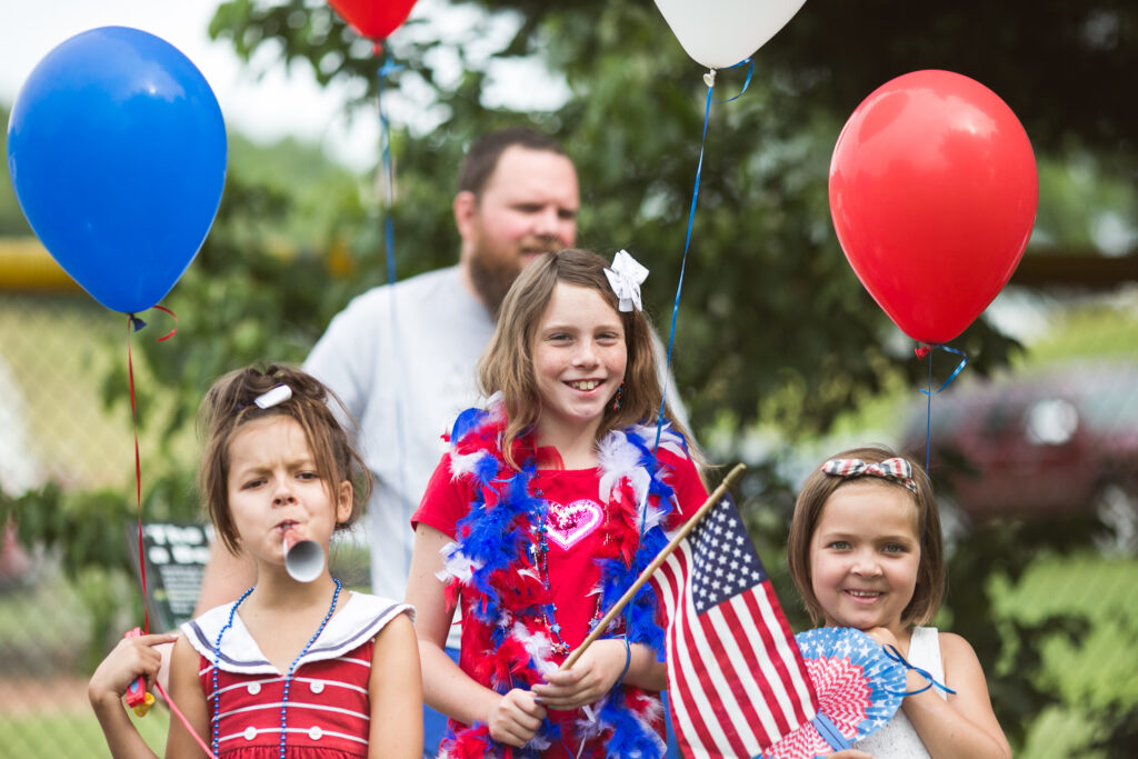 Children in red white and blue patriotic outfits holding flags and balloons.