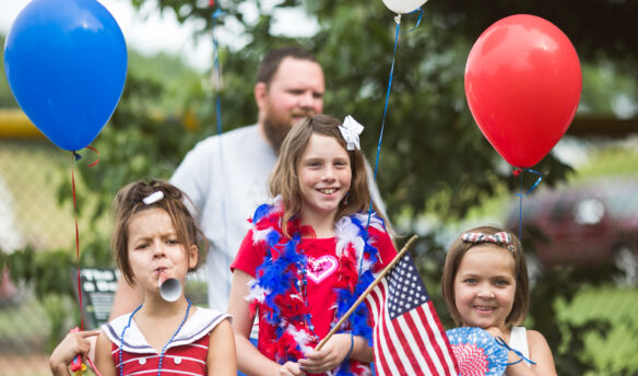 Children in red white and blue patriotic outfits holding flags and balloons.